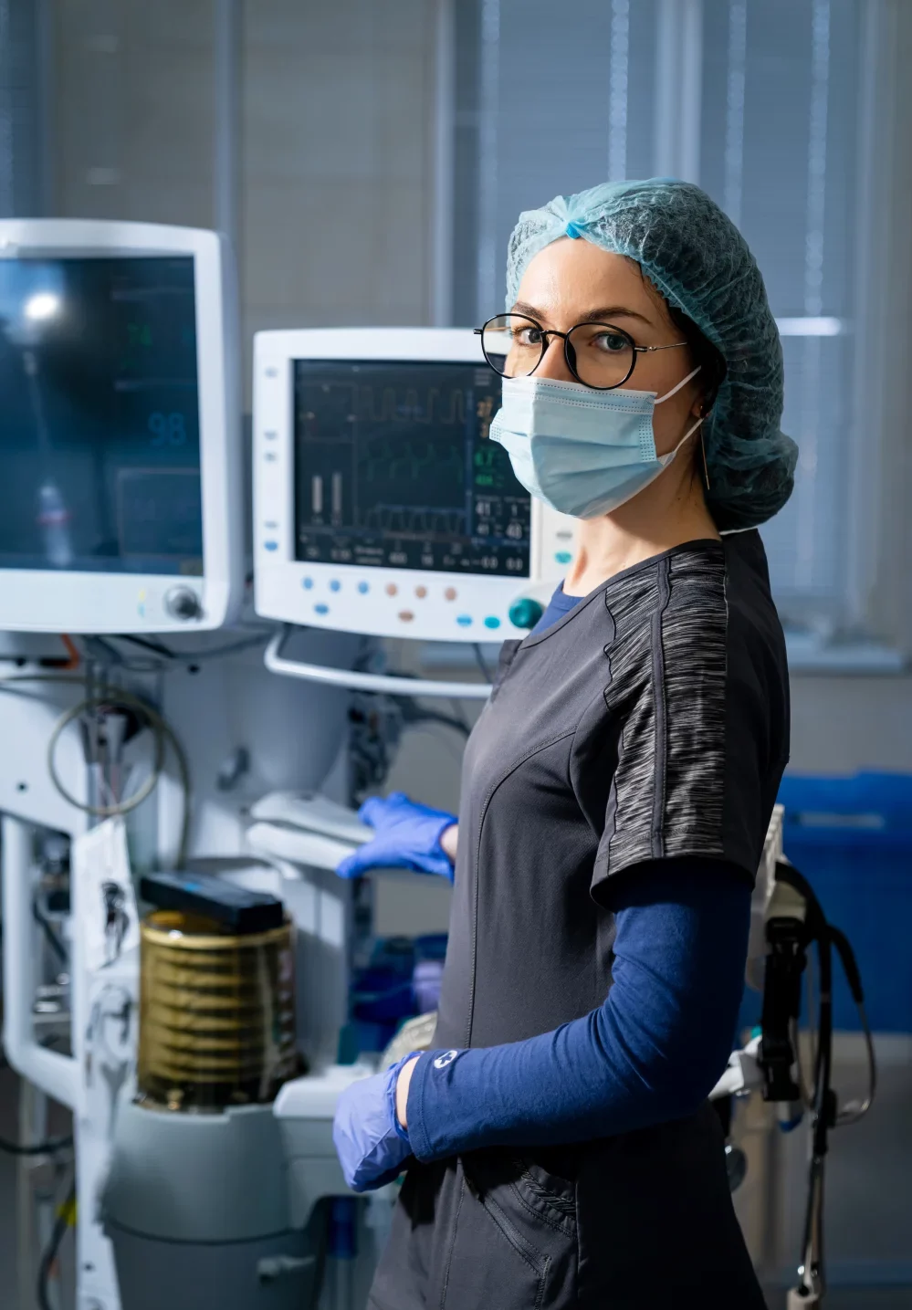 Medical professional in a mask and scrub cap standing beside patient monitoring equipment
